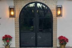 This image features a stylish arched glass door framed by decorative tiles and illuminated by outdoor wall sconces, enhancing the building's curb appe.
