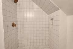 A contemporary shower area featuring white tiles, wooden fixtures, and a sloped ceiling, ideal for modern bathroom designs.
