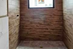 This image showcases a bathroom under construction featuring wood-textured wall tiles, a small window, and a tiled floor.