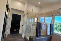Bright bathroom with glass shower, black marble base, and tropical view through large windows.